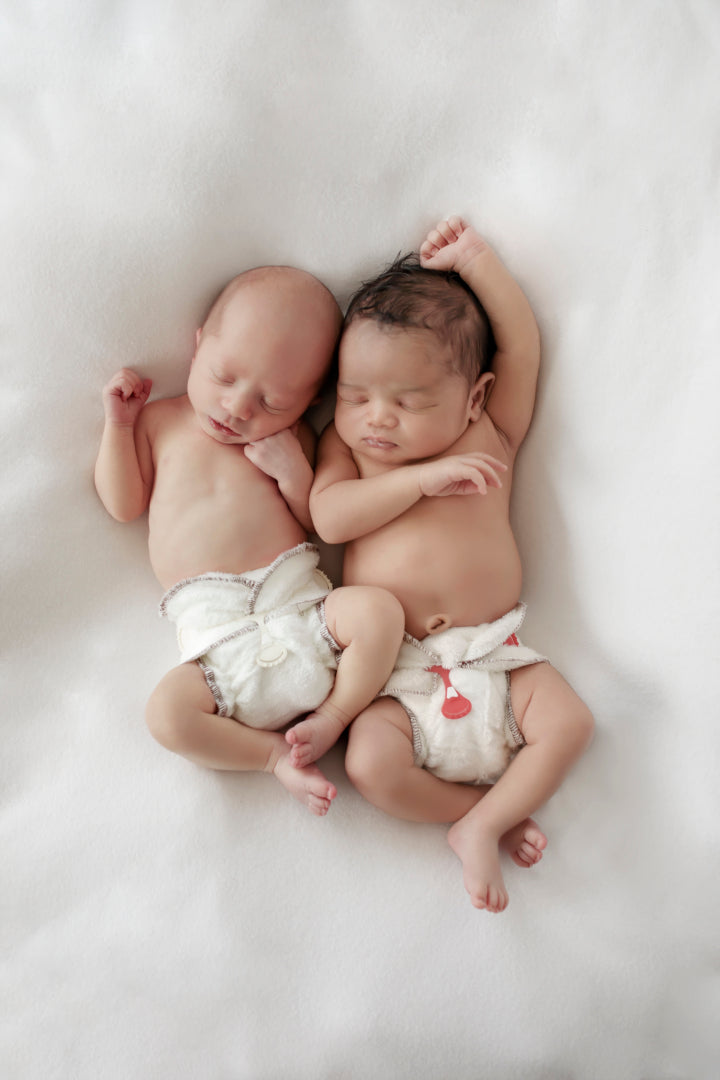 Two sleeping babies are wearing white Bubblebubs Bambam cloth nappies with a white snappi and a red snappi with their hands over their heads.