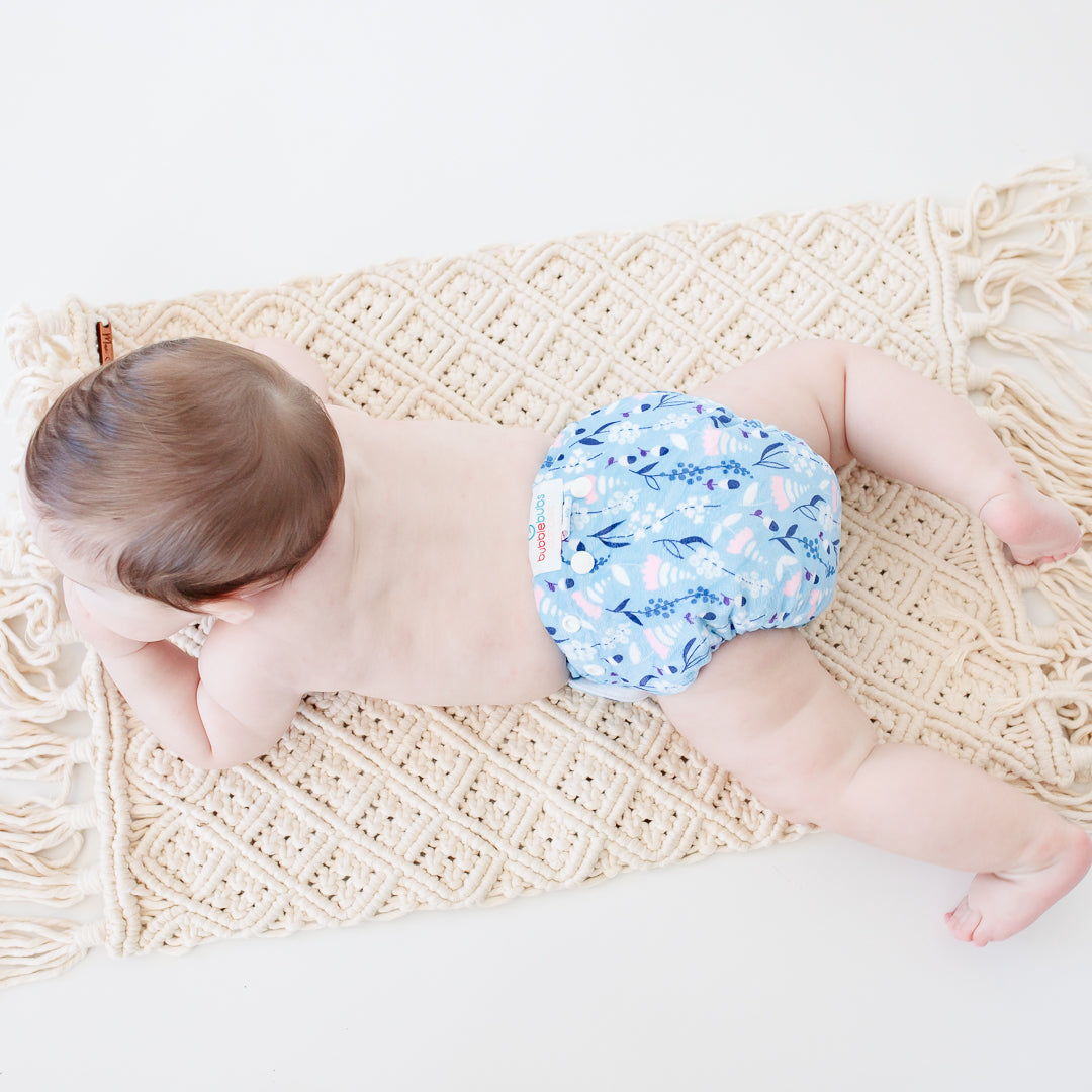 Baby lying on a patterned mat wearing a Bubblebubs Candie with a blue, white and pink pattern. The back of the baby is facing the camera, but you can see the Candie cloth nappy fits so well that it does not leak.