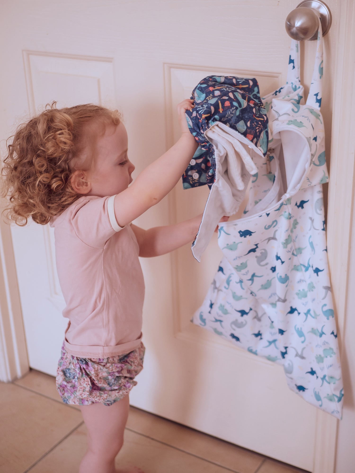 The toddler puts used cloth nappies into an XL wet bag hanging on the doorknob. She has curly red hair and is wearing pink clothes.