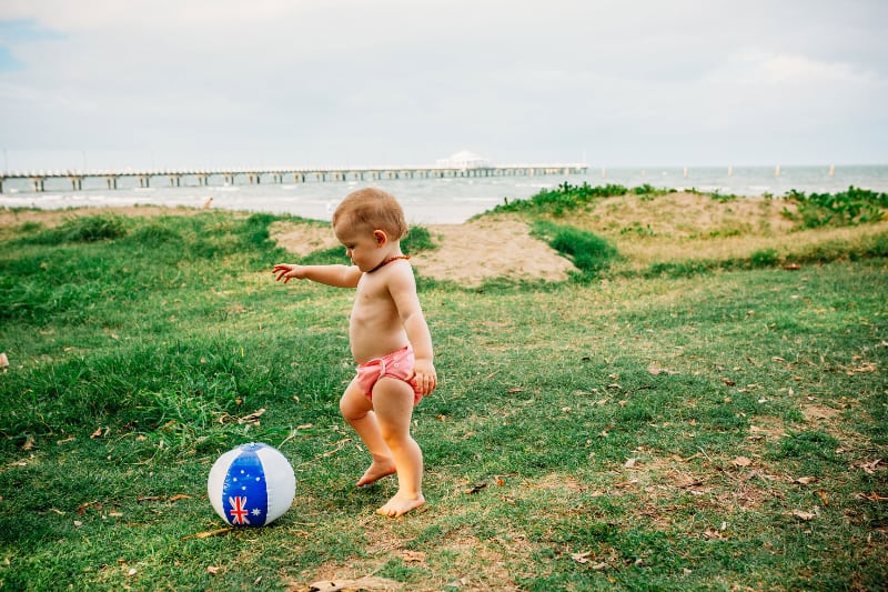 Toddler chasing a beachball with the Australian flag in a peach-coloured Bubblebubs Candie cloth nappy on a beach.