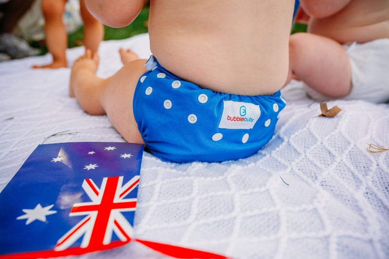 Baby lying on a white blanket with an Australian flag waring a blue Bubblebubs Candie cloth nappy.