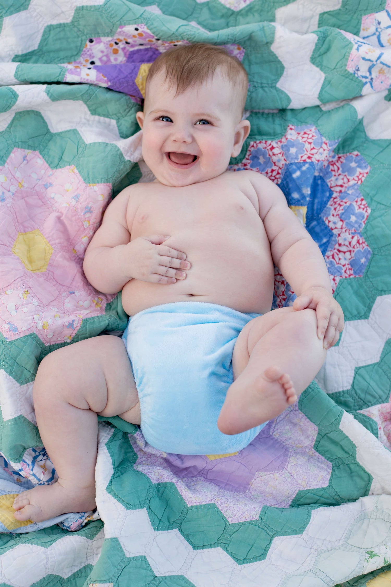 A very happy toddler is lying on a blanket, showing off the blue Bubblebubs Candie cloth nappy he is wearing. 
