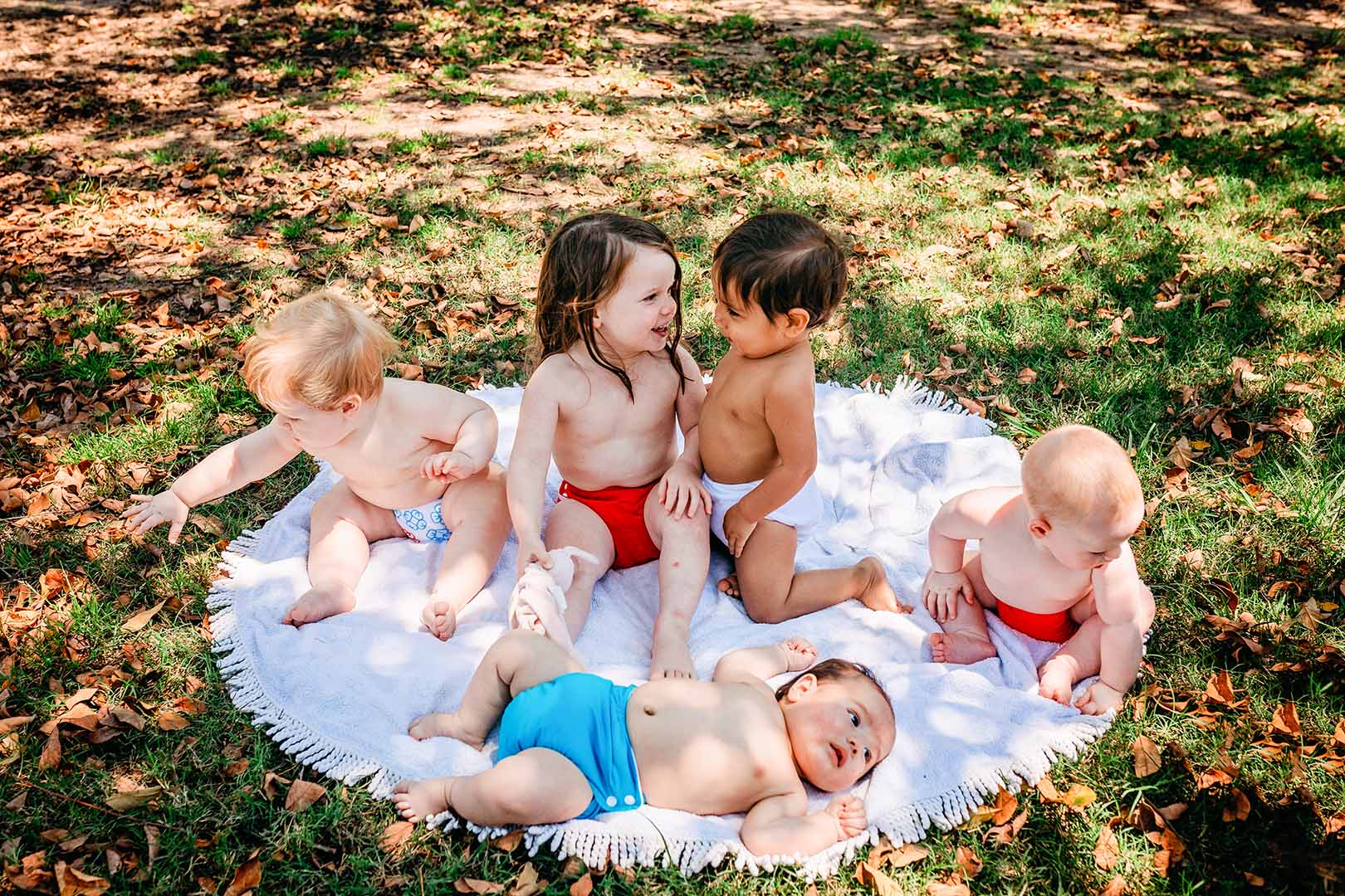 5 babies playing outside in the shade of a tree. Each has a different colour cloth nappy on, and they fit all the babies snugly so no leaks can happen—Blue, red and white reusable cloth nappies