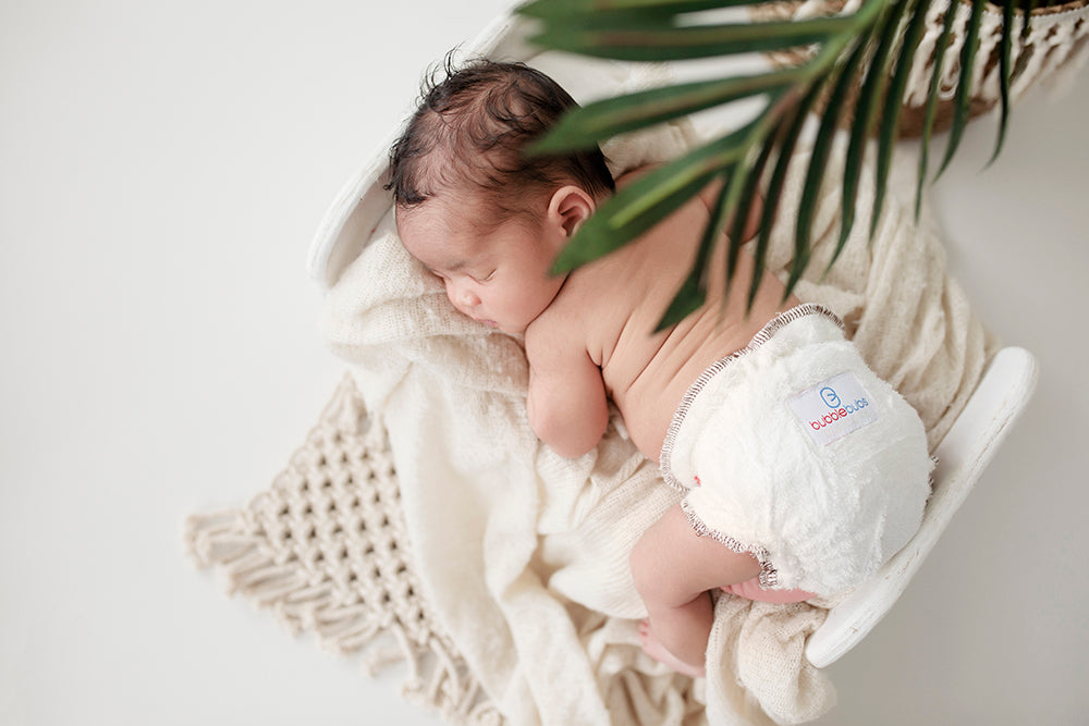 Dark haired baby sleeping in a good fitting Bubblebubs Bambam cloth nappy under the shade of a palm tree on different white blankets.
