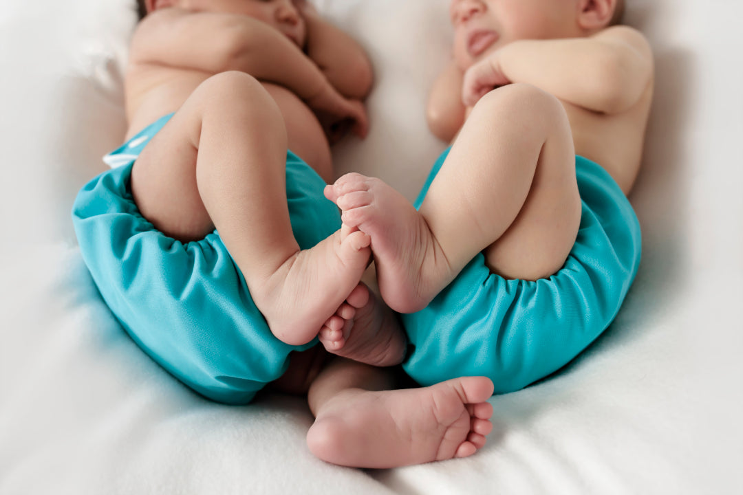 Two babies touching feel as they sleep in matching blue Bubblebubs Candie cloth nappies. They look like they could be twins with their matching reusable nappy.