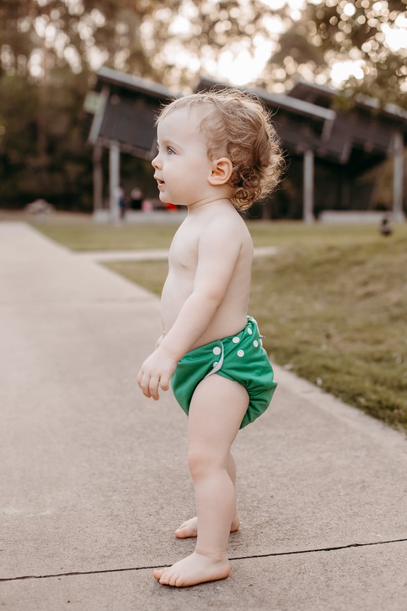 A toddler is standing on a path looking to the side, waring a green Bubblebubs Candie cloth nappy.